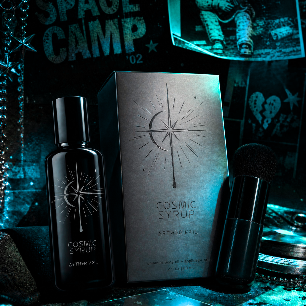 Cosmic Syrup Aether Veil shimmering dry body oil bottle and packaging on a reflective surface with 'Space Camp' text in the background